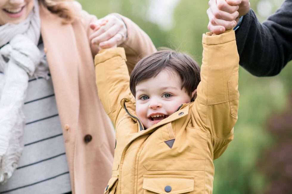 Toddler swinging between parents' arms during a family portrait session in the park near Ilkley, Yorkshire