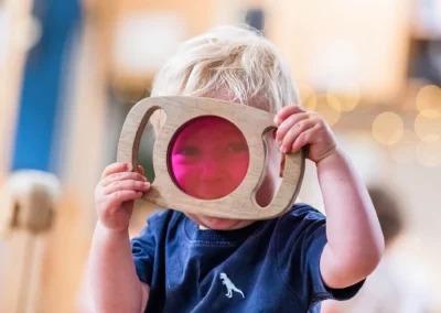 Young child holding a wooden magnifying glass during a forest school session, photographed for nursery marketing in Leeds, West Yorkshire
