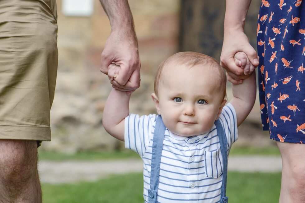 Baby standing with parents' support during an outdoor family photography session in Skipton, North Yorkshire