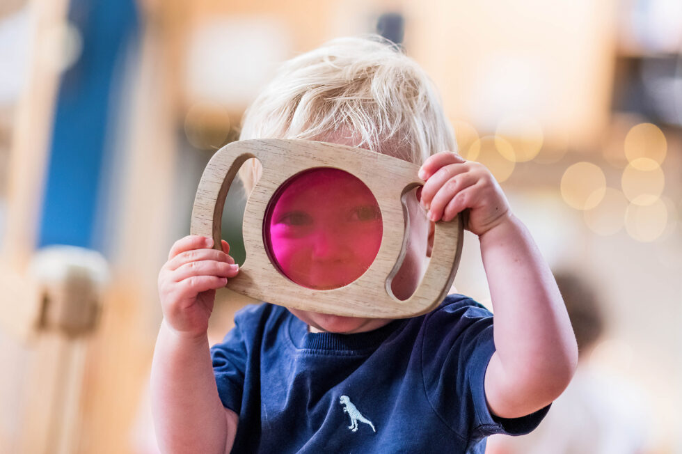 Young child holding a wooden magnifying glass during a forest school session, photographed for nursery marketing in Leeds, West Yorkshire